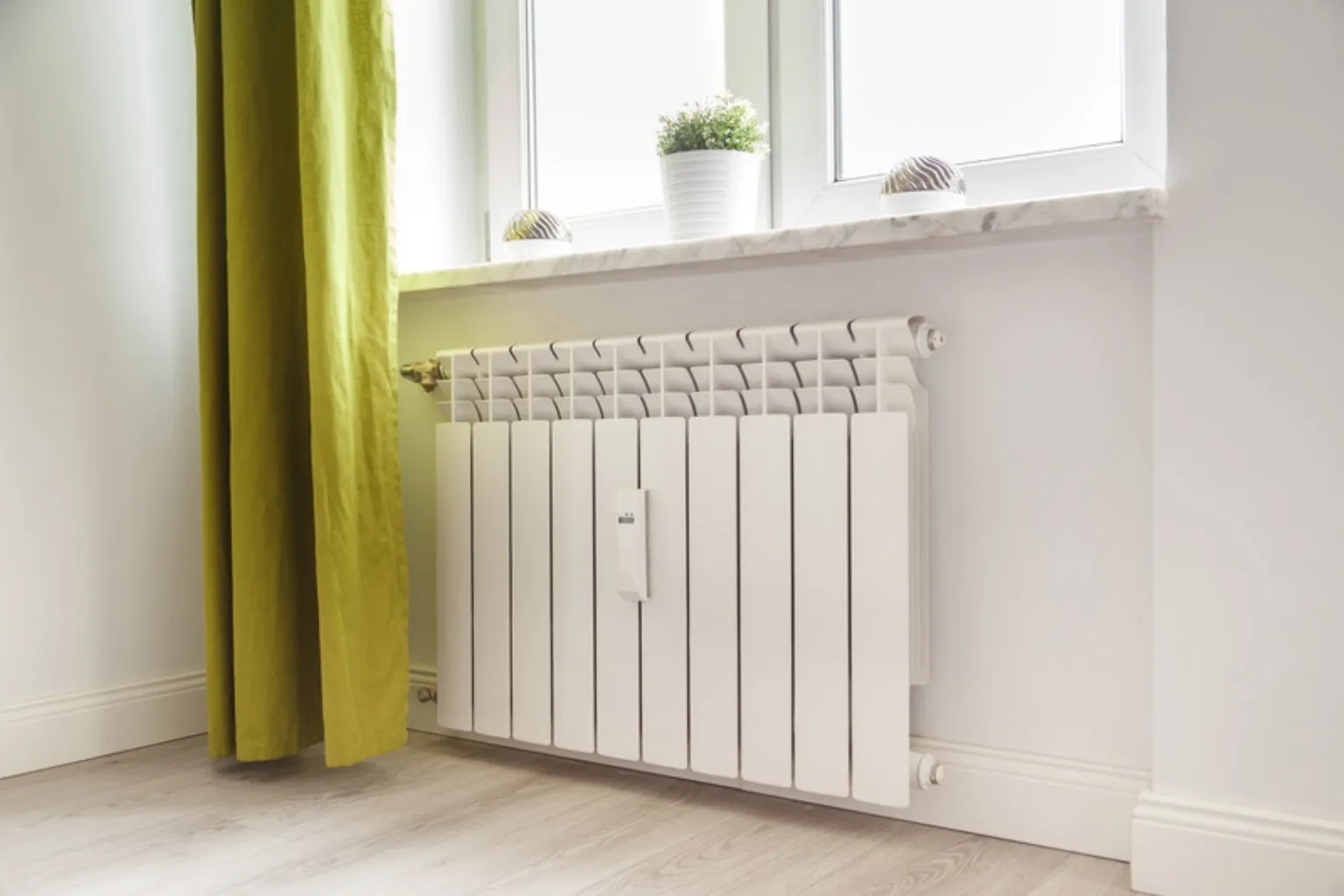 Hvid radiator under et vindue i en lys stue med gardiner